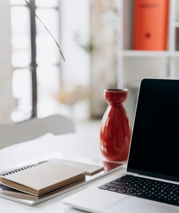 A minimalist home office setup featuring a laptop, notebook, and a red vase on a white desk.